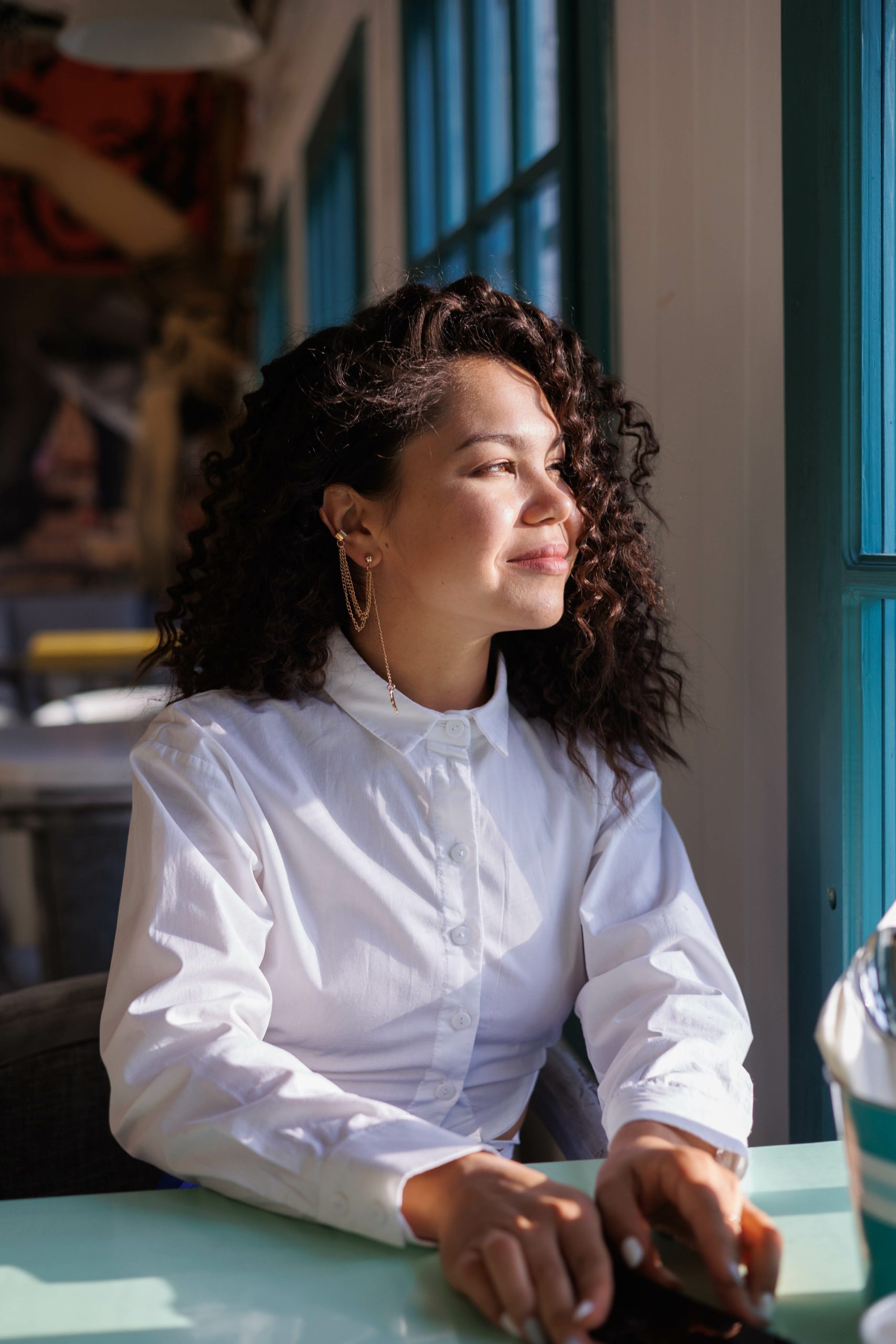Happy curly brunette girl smiling in cafe. Young woman enyoing sun in restaurant at window. white blouse. Sun in city. Fashionable asian girl with frizzly hair. Sun light in window